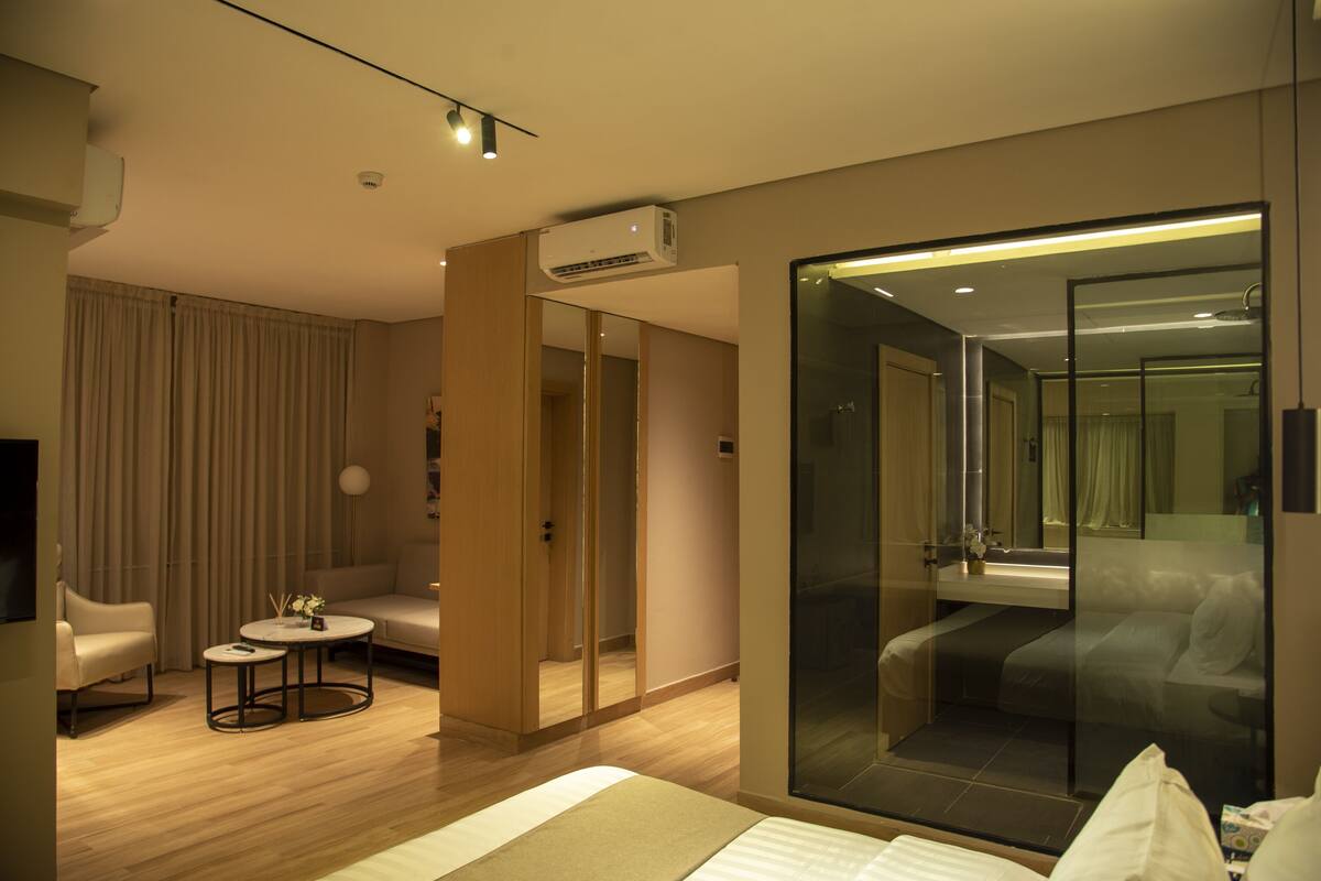 A modern hotel room with a bed, a seating area with a small table, a glass-enclosed shower, and a large mirror.