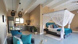A modern bedroom with a canopy bed, a sitting area with a sofa, and a kitchen in the background. - Marepe Mountain View (Hoedspruit)