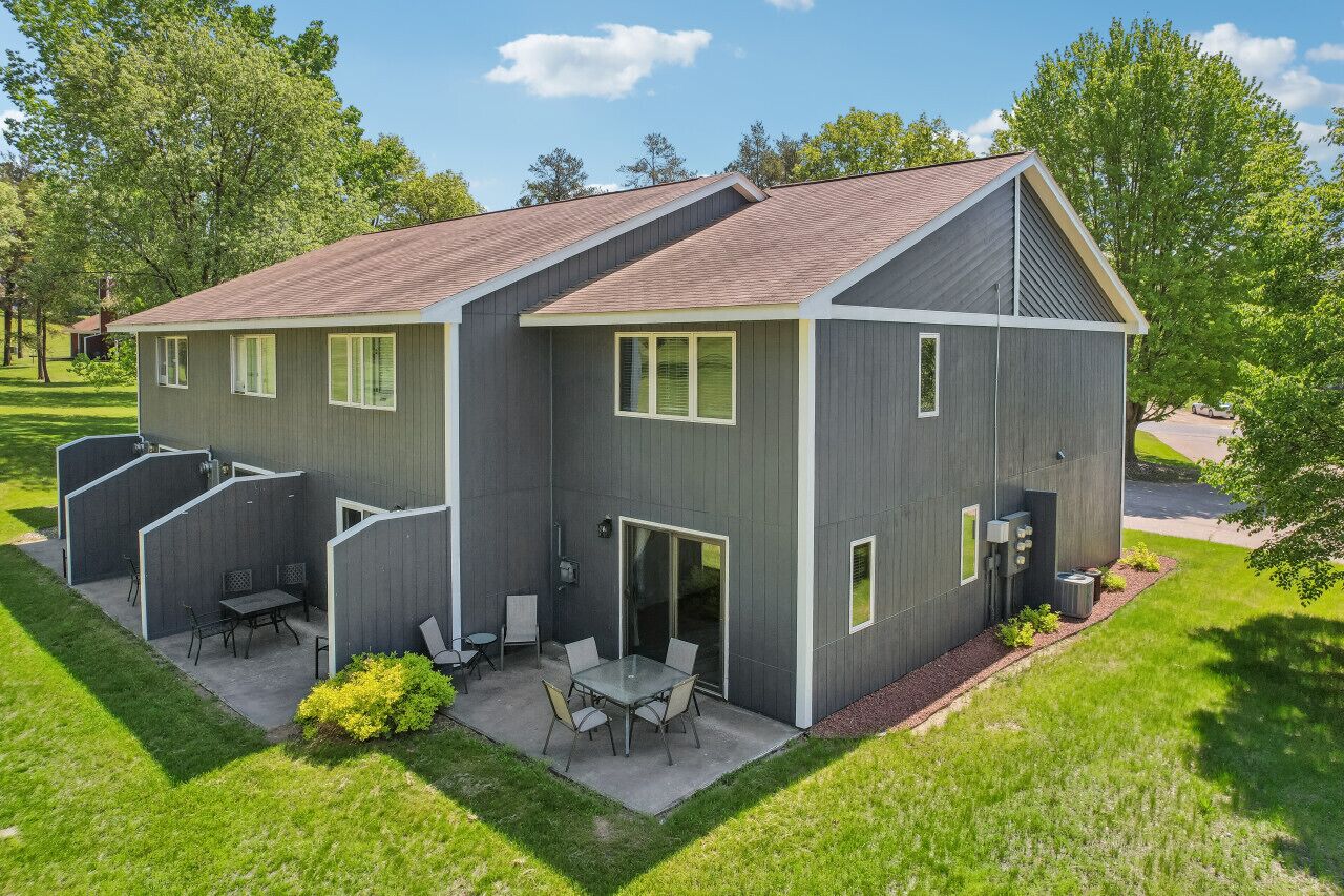 Un bâtiment moderne à deux étages, doté d’un toit plat, d’un patio couvert avec une table et des chaises, et d’un petit jardin.