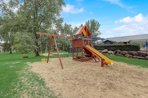 Un parque infantil con columpios y un tobogán, rodeado de césped y árboles.