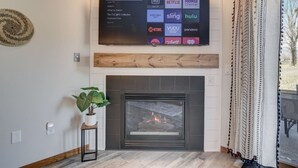 Un téléviseur à écran plat fixé au-dessus d’une cheminée où l’on voit le feu brûler, une plante en pot et un thermostat fixé au mur.