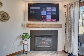Un téléviseur à écran plat fixé au-dessus d’une cheminée où l’on voit le feu brûler, une plante en pot et un thermostat fixé au mur.