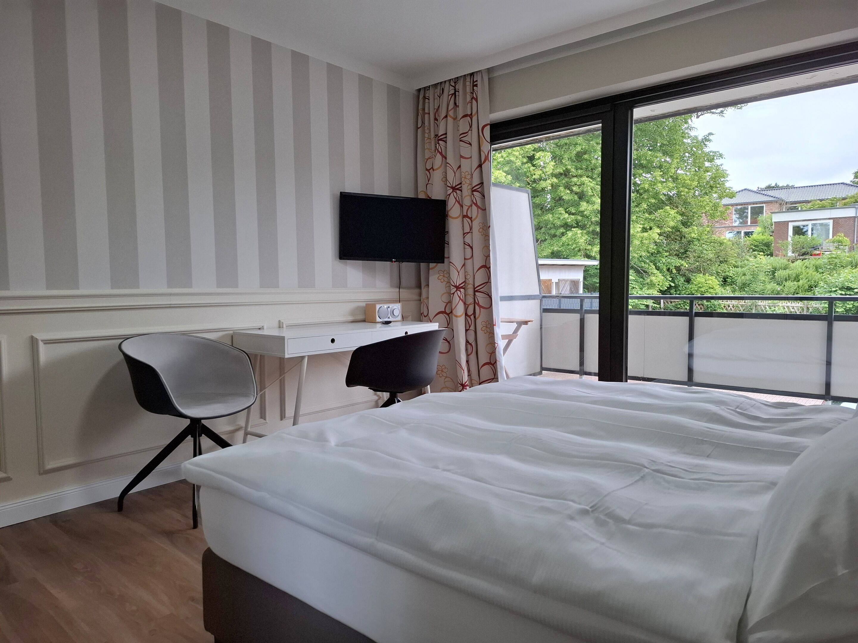 Hotelkamer met een bed, bureau, stoel, televisie en een balkon met uitzicht op gebouwen en bomen.