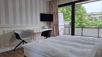 Hotelkamer met een bed, bureau, stoel, televisie en een balkon met uitzicht op gebouwen en bomen.