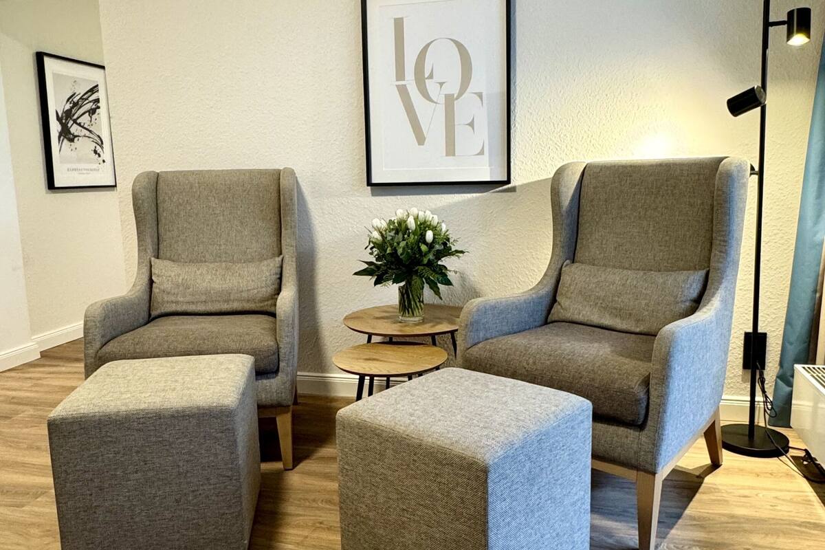 Una sala de estar moderna con dos sillones grises, un reposapiés a juego, una pequeña mesa redonda con una planta y un cartel en la pared con la palabra 'LOVE'.