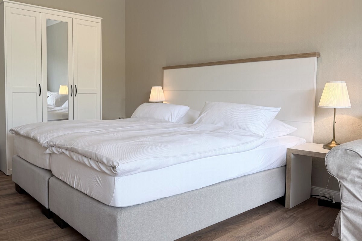 Una cama bien hecha con sábanas blancas, un cabecero de madera, una mesita de noche con una lámpara y un amplio armario con puertas espejadas.