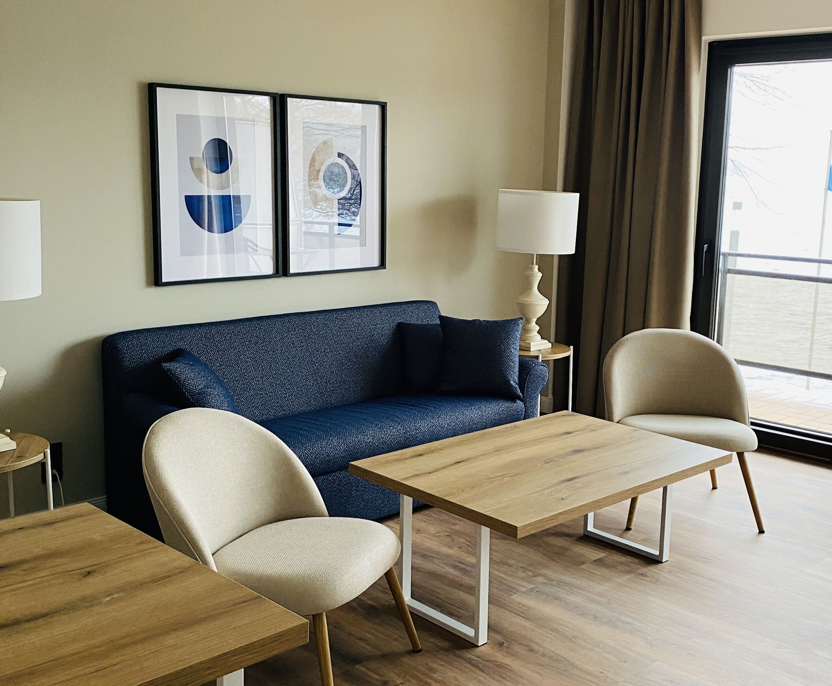 Habitación de hotel moderna con un sofá azul, dos mesas de madera, un sillón color beige y dos obras de arte abstractas enmarcadas en la pared.