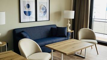 Habitación de hotel moderna con un sofá azul, dos mesas de madera, un sillón color beige y dos obras de arte abstractas enmarcadas en la pared.