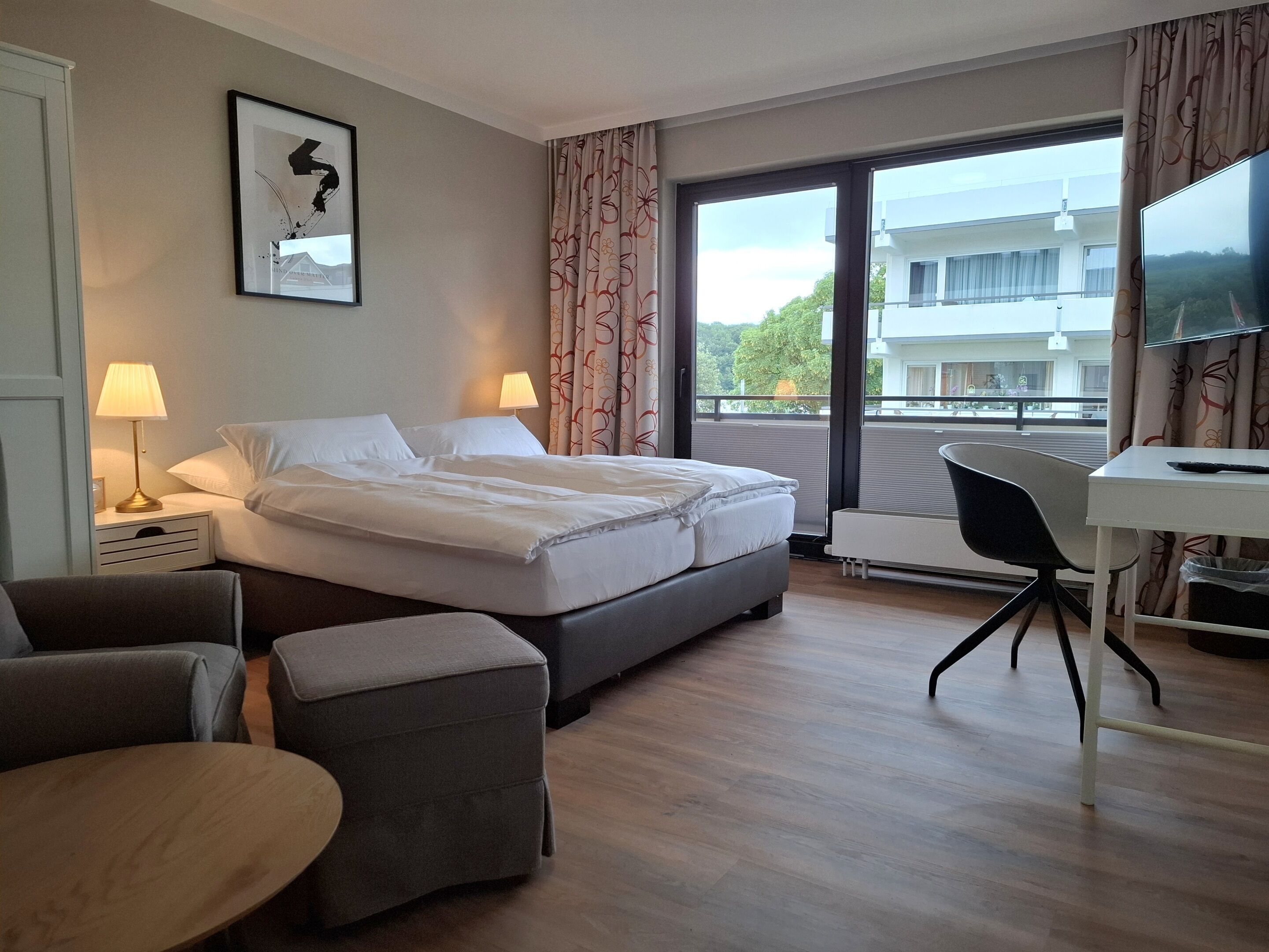 Habitación de hotel con cama, lámparas de noche, escritorio, silla y un amplio ventanal con vista a edificios.