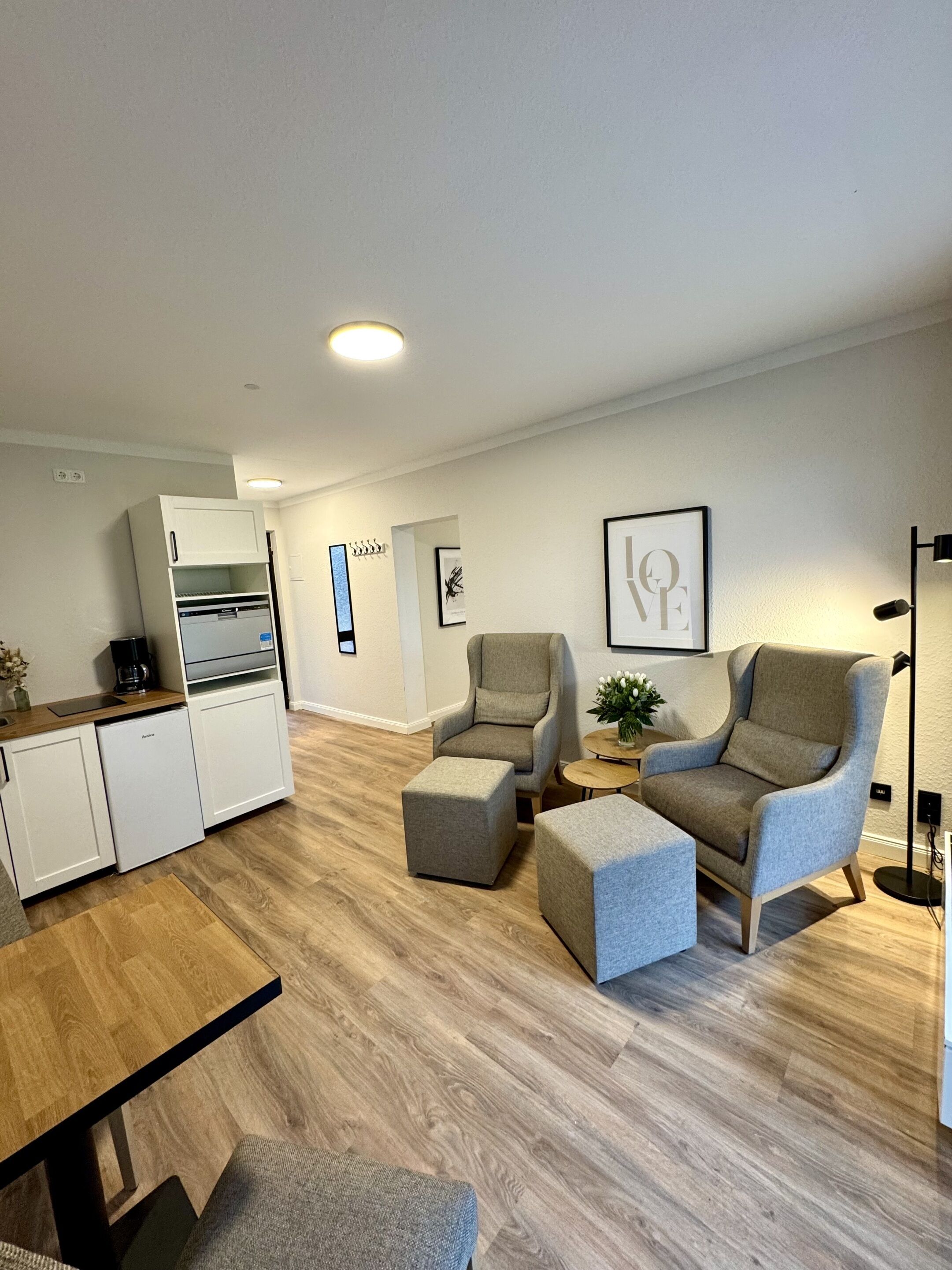 Ein modernes Wohnzimmer mit Sofa, Hocker und einem kleinen Tisch mit Pflanze. Es gibt eine Küchenzeile mit Kühlschrank und Kaffeemaschine.