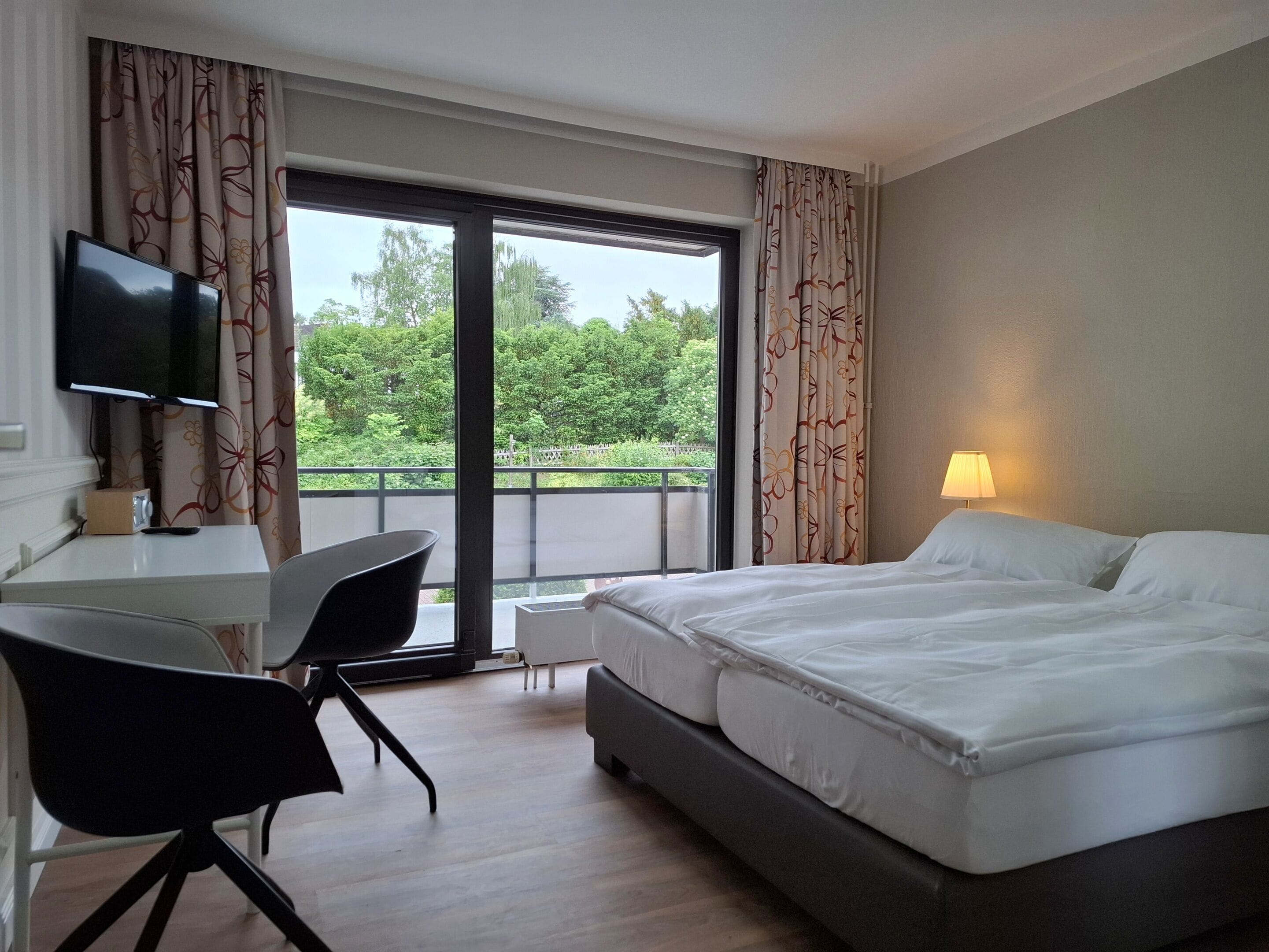 Ein Hotelzimmer mit Bett, Schreibtisch mit Stuhl, Fernseher und einem Balkon mit Vorhängen.