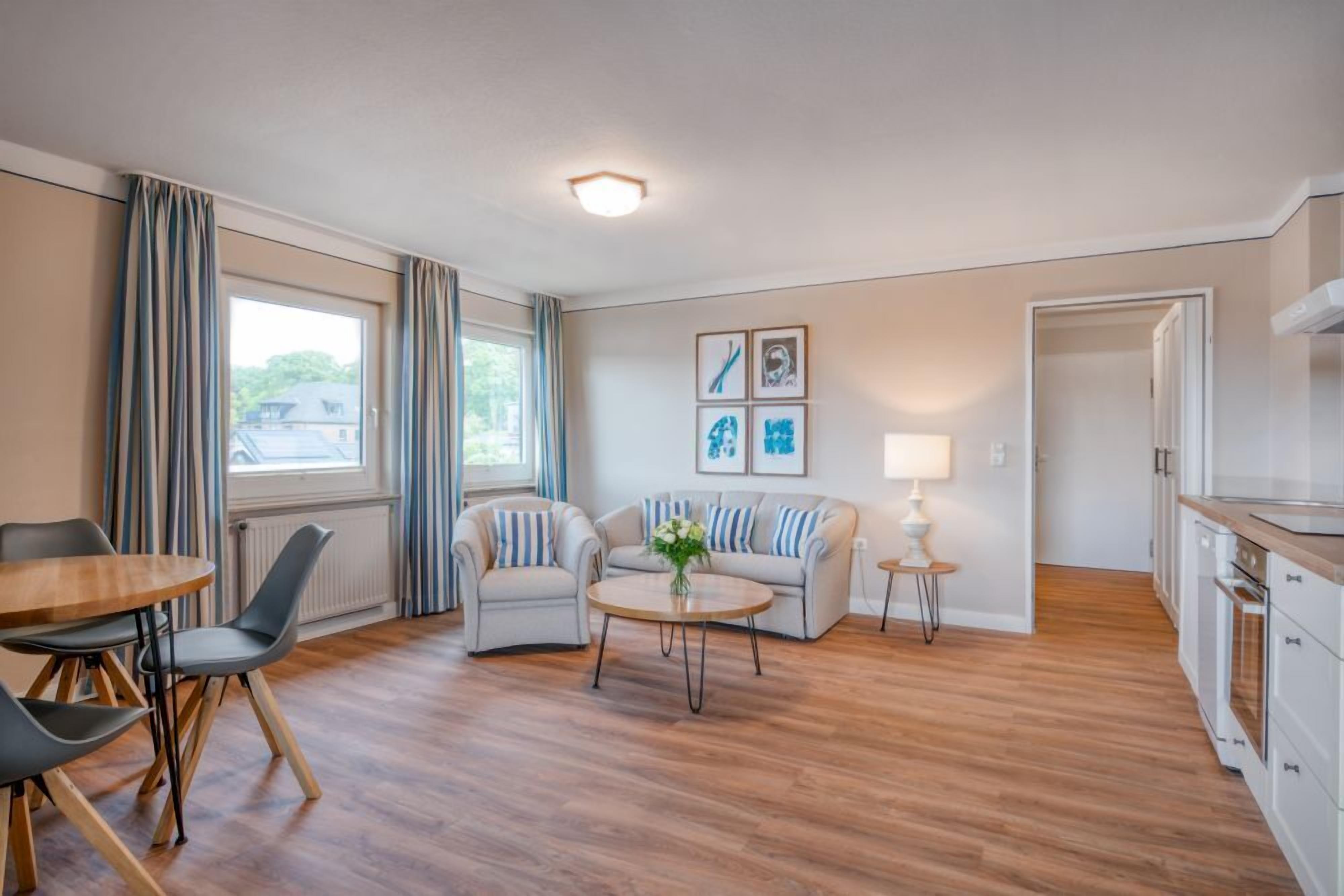 Sala moderna com piso de madeira, sofá, mesa de centro e área de jantar com cadeiras. Há também uma cozinha com armários e uma geladeira.