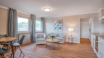 Sala moderna com piso de madeira, sofá, mesa de centro e área de jantar com cadeiras. Há também uma cozinha com armários e uma geladeira.