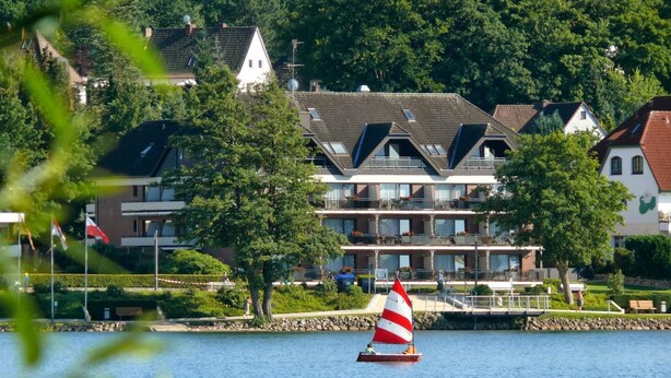 A sailboat on a calm lake with a row of houses in the background. - Landhuus Dieksee (Malente)