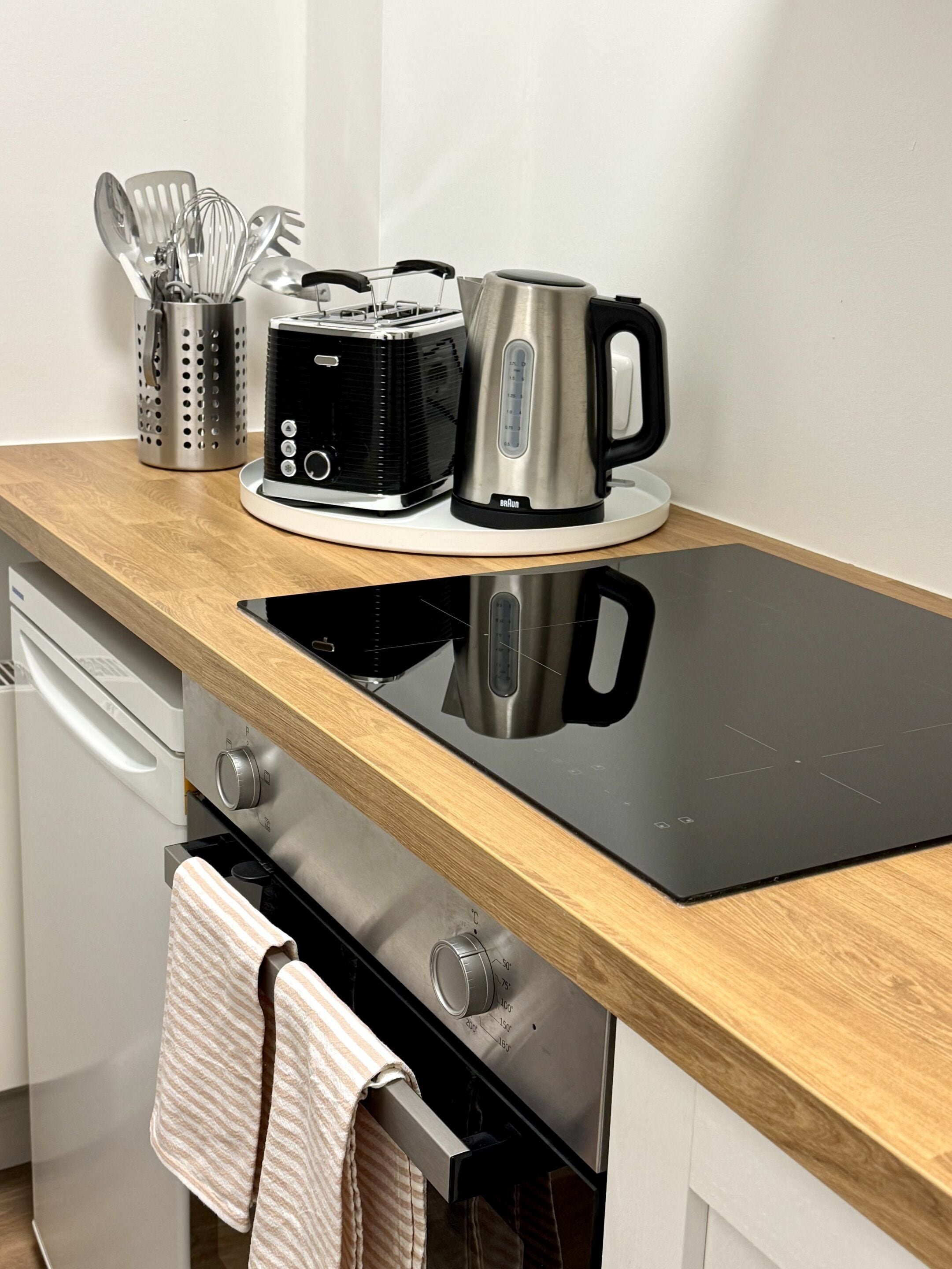 Una cocina con tope de inducción negro, una tetera, un tostador y un juego de utensilios sobre la encimera.