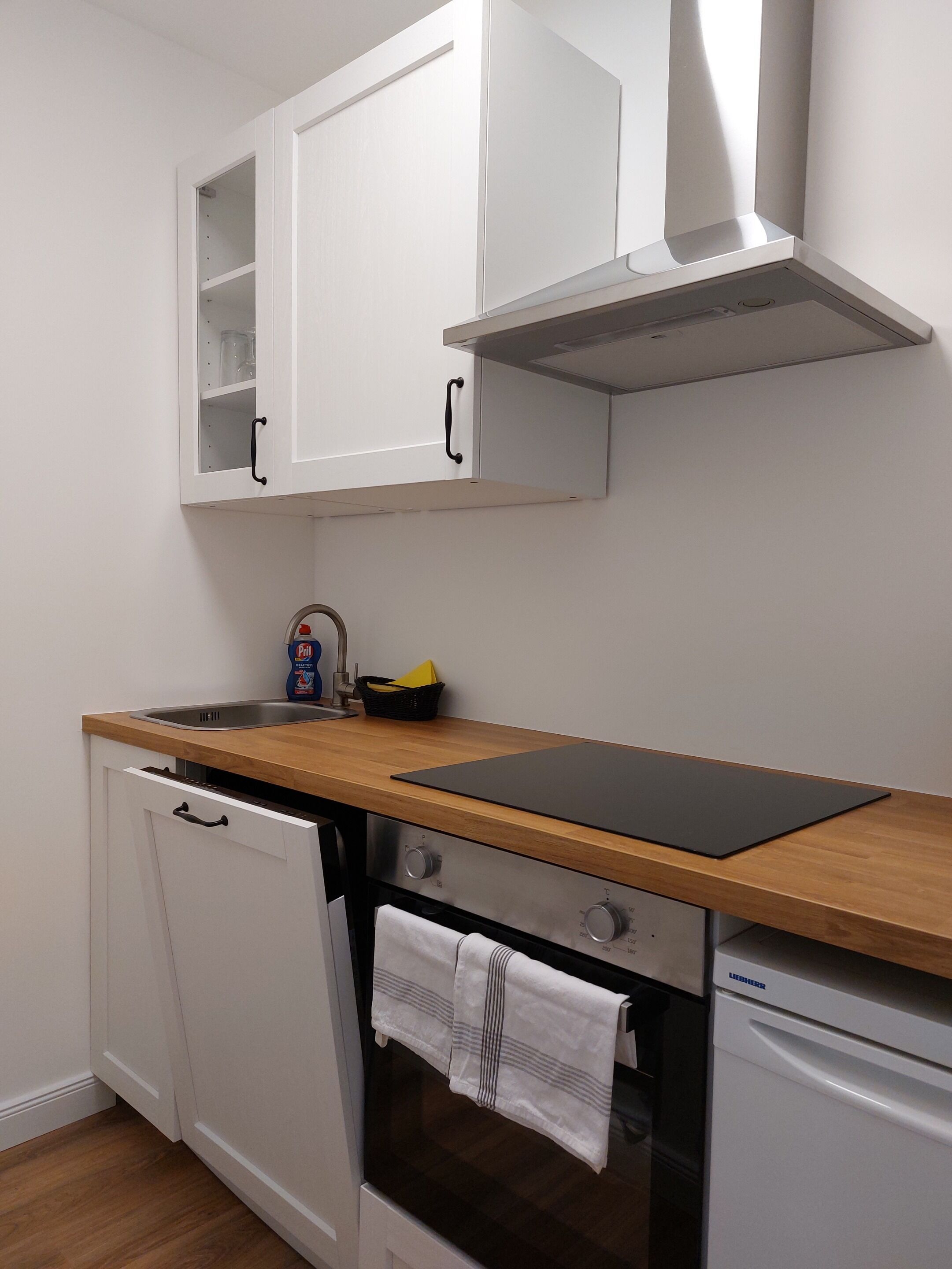 A compact kitchen with white cabinets, a wooden countertop, a built-in oven, and a dishwasher.