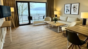 A modern hotel room with a wooden floor, a sofa, a dining table, a TV, and a balcony with a view of a lake. - Landhuus Dieksee (Malente)