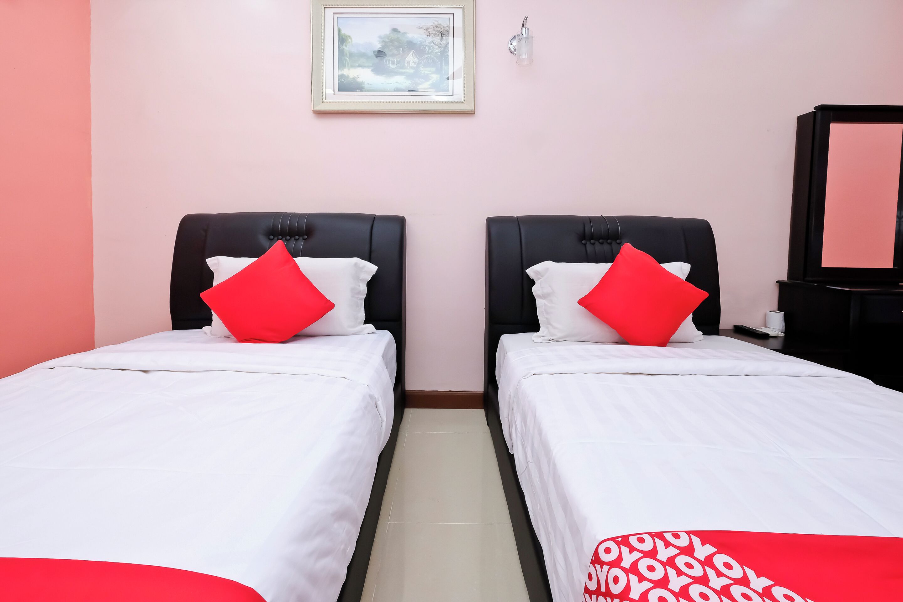 Habitación de hotel con dos camas, cada una con ropa blanca y almohadas rojas. Se observa un cuadro en la pared y un mueble negro a la derecha.