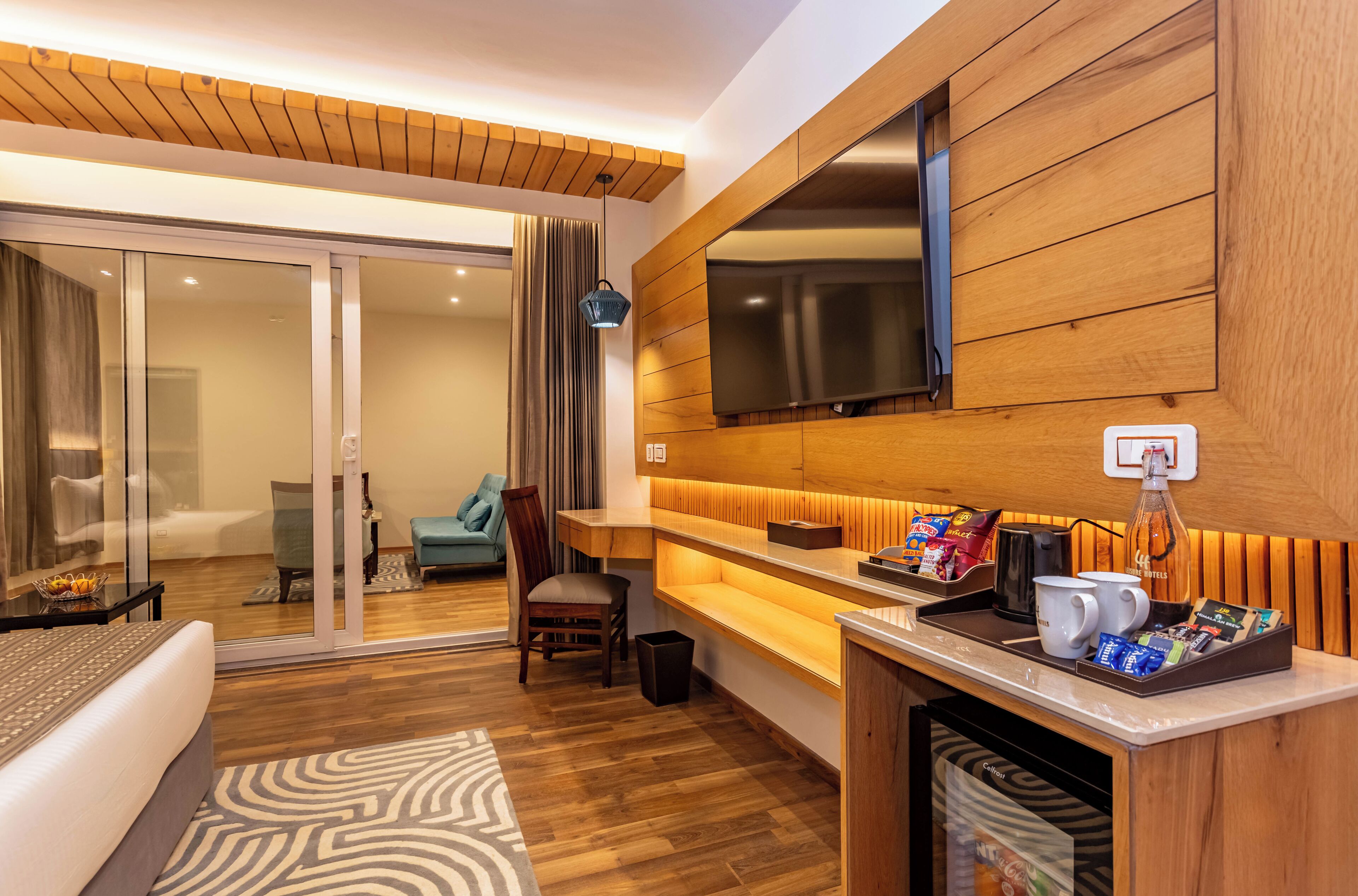 A hotel room with a wooden floor, a sofa, a chair, a TV mounted on the wall, a coffee station, and a sliding glass door leading to a balcony.