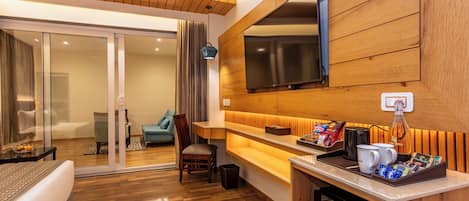 A hotel room with a wooden floor, a sofa, a chair, a TV mounted on the wall, a coffee station, and a sliding glass door leading to a balcony.