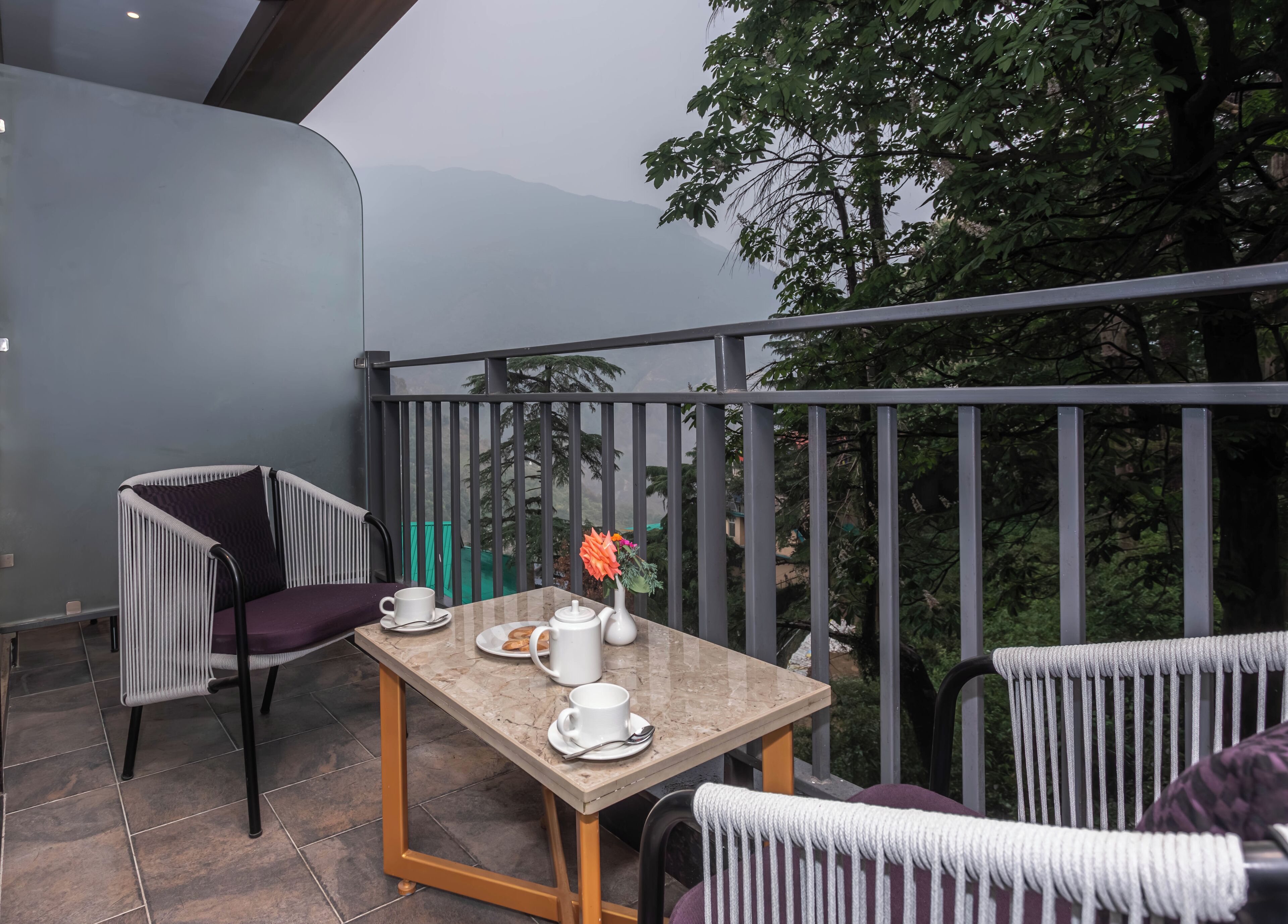 A balcony with a table set for two, featuring a white teapot, cups, and a vase with flowers.