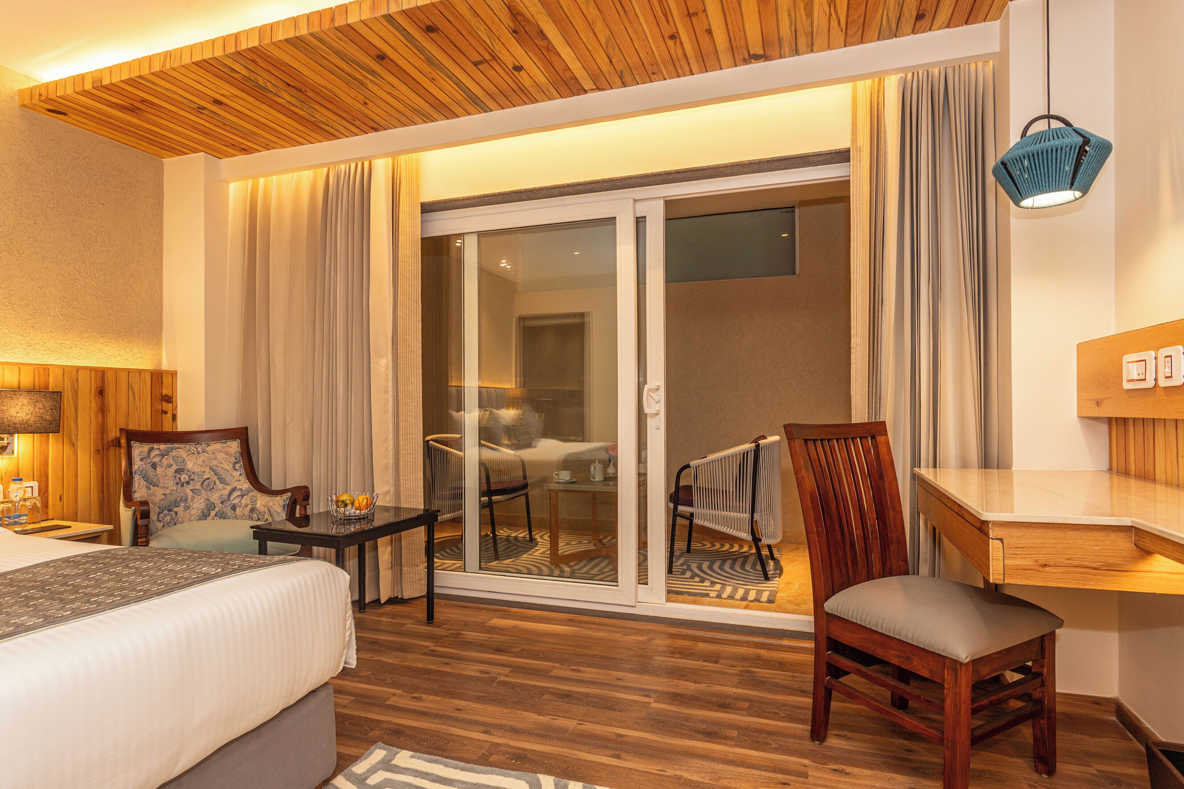 Une chambre d’hôtel avec un plafond en bois, un lit, un bureau, une chaise, une petite table avec un panier, et une porte-fenêtre coulissante donnant sur un balcon.