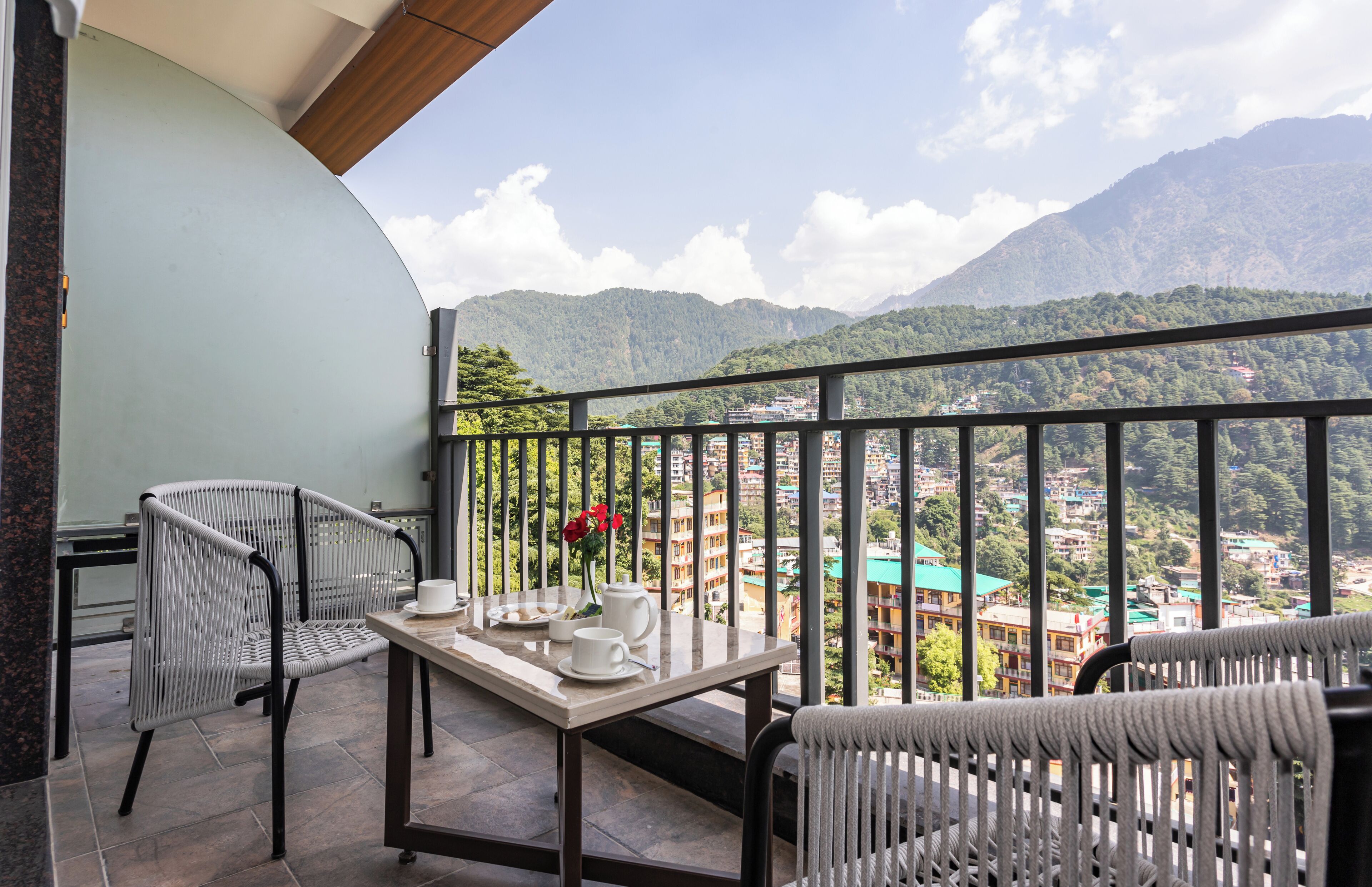A balcony with a table set for tea, two wicker chairs, and a view of a mountainous town.