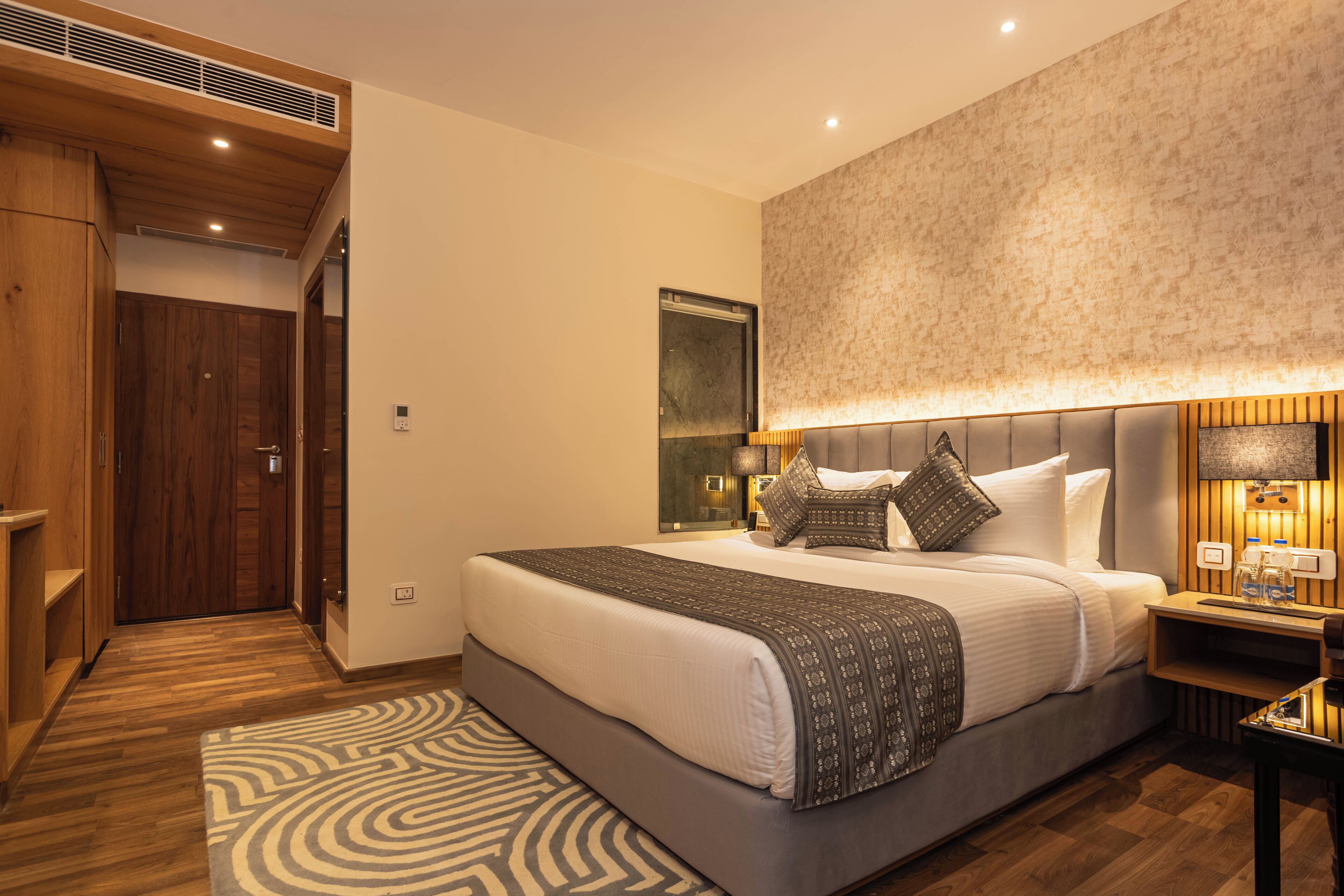 A modern hotel room with a large bed, wooden flooring, a patterned rug, and a built-in wardrobe.