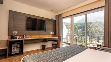 A hotel room with a large bed, a flat-screen TV mounted on the wall, a mini-fridge, a small table with a chair, and a balcony with a mountain view.