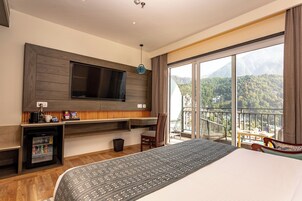 A hotel room with a large bed, a flat-screen TV mounted on the wall, a mini-fridge, a small table with a chair, and a balcony with a mountain view.