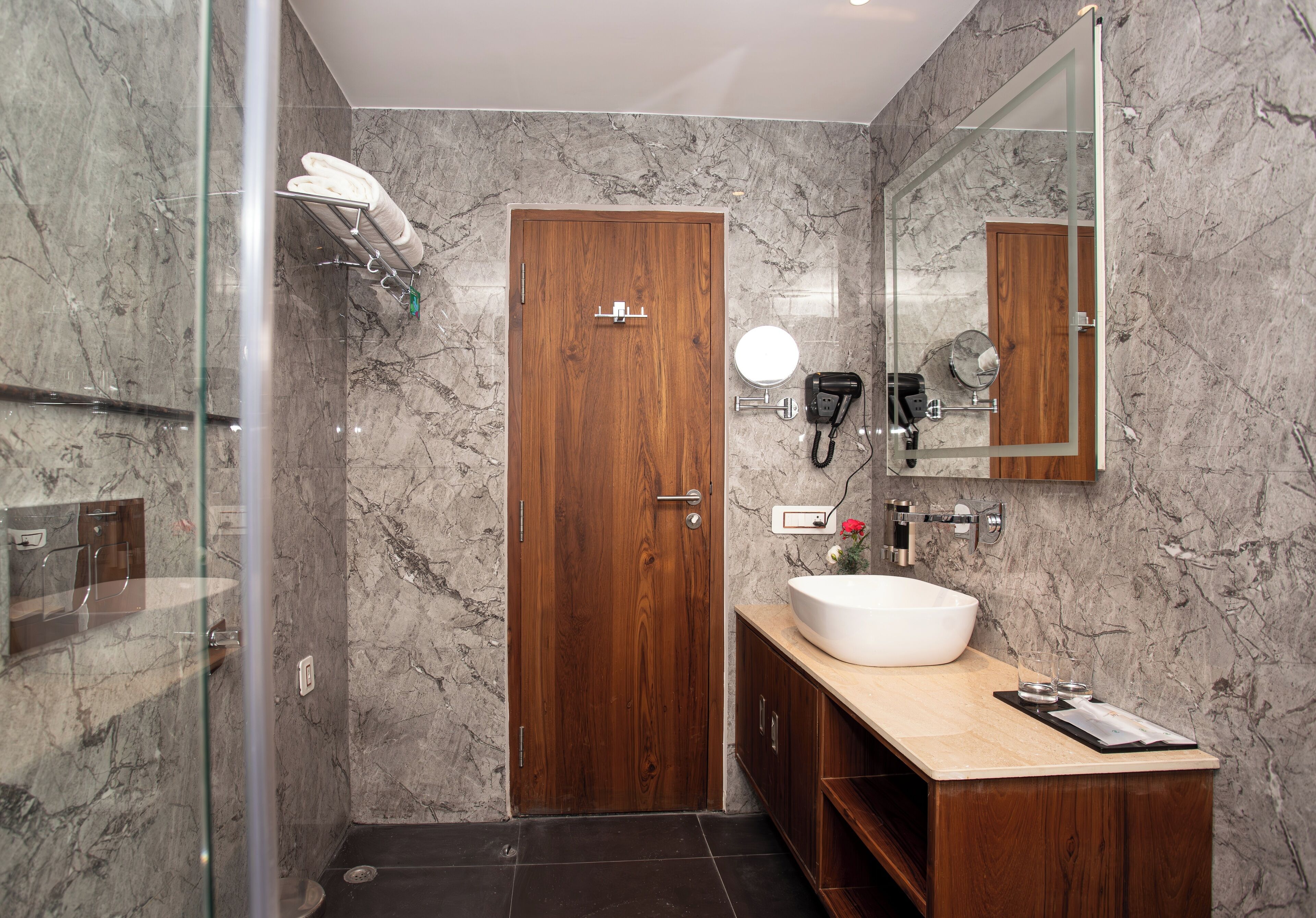 Un baño moderno con puerta de madera, cabina de ducha de vidrio, pared de mármol, tocador con un lavamanos grande y un espejo.