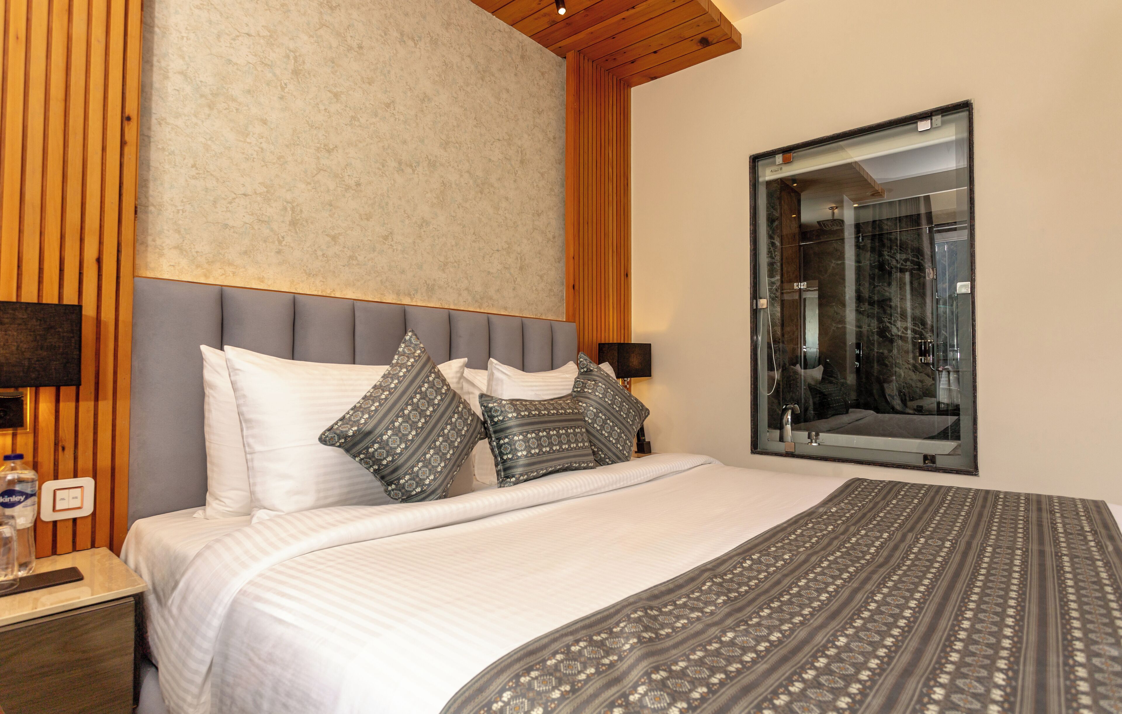 A bedroom with a large bed, a wooden headboard, a mirror, and a nightstand with a lamp and a bottle.