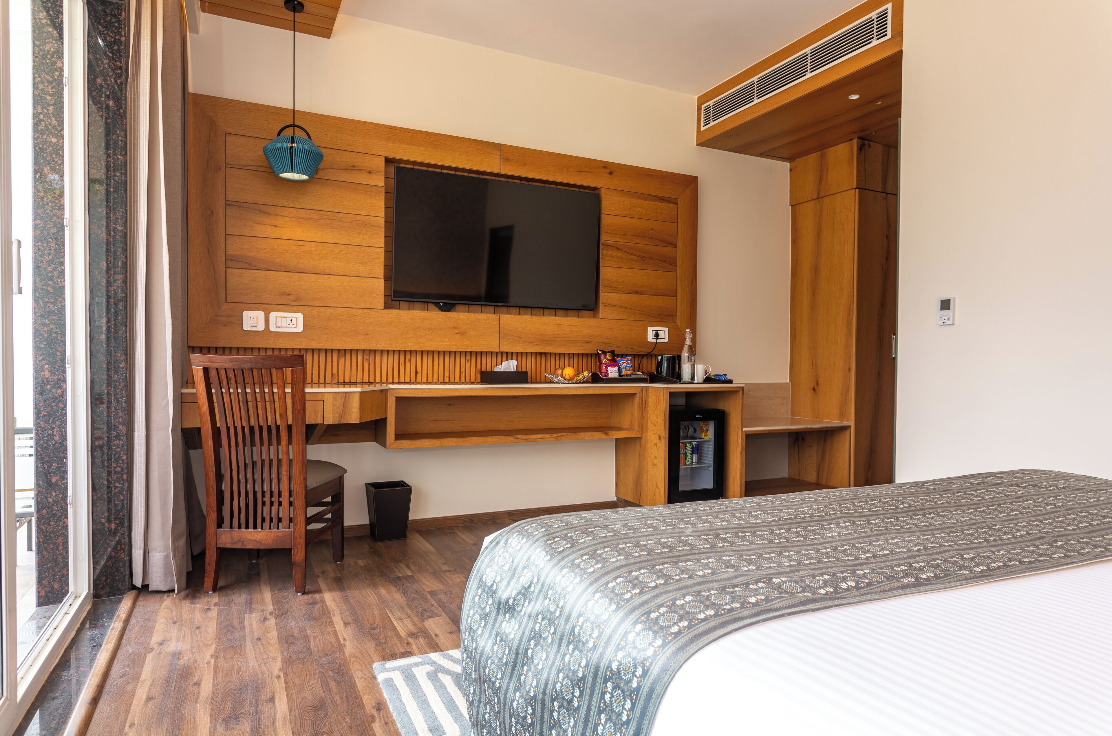 A bedroom with a bed, a TV mounted on a wooden wall, a desk with a chair, and a small refrigerator.
