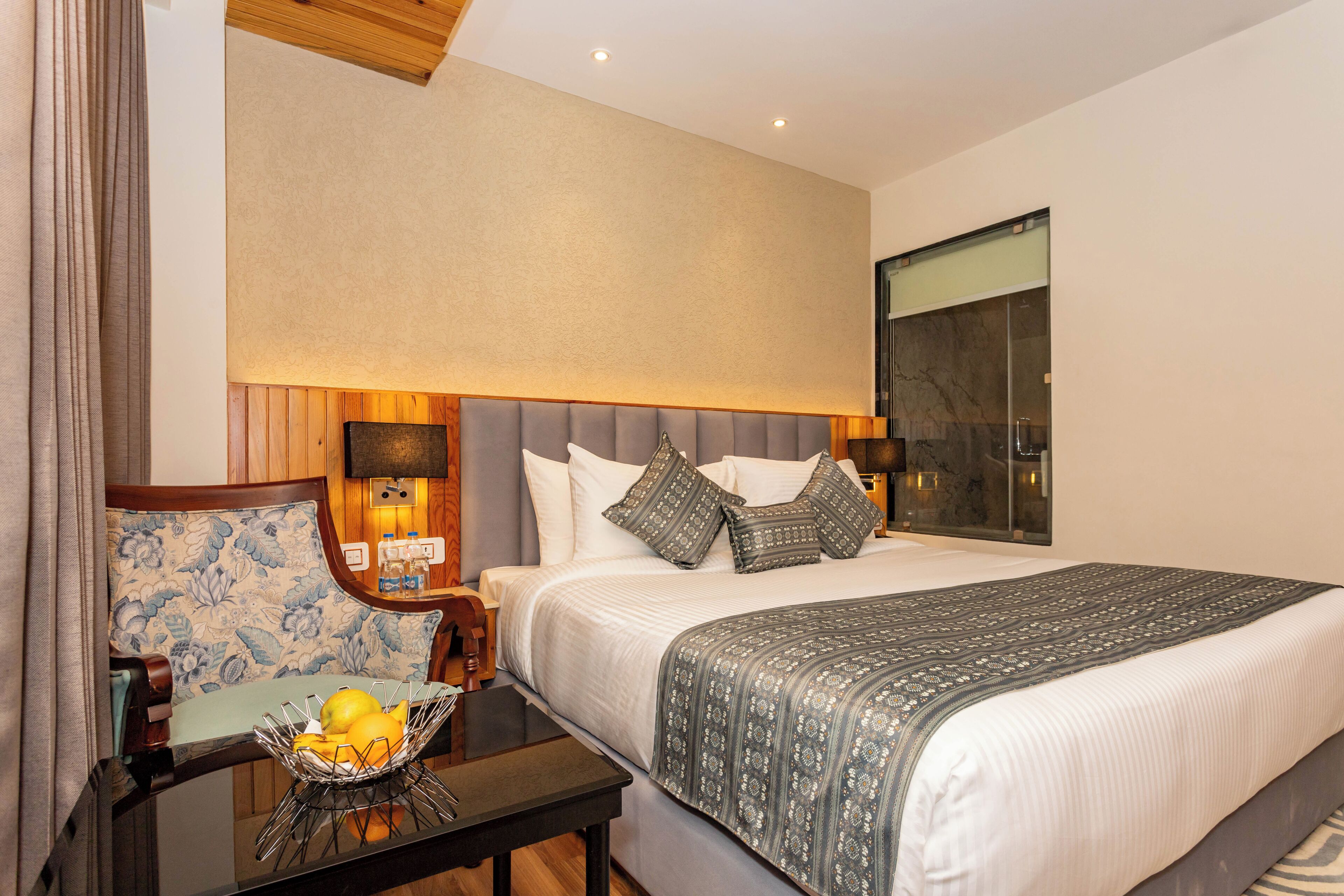 A hotel room with a large bed, a bedside table with a fruit bowl, a chair with a floral cushion, and a large window.
