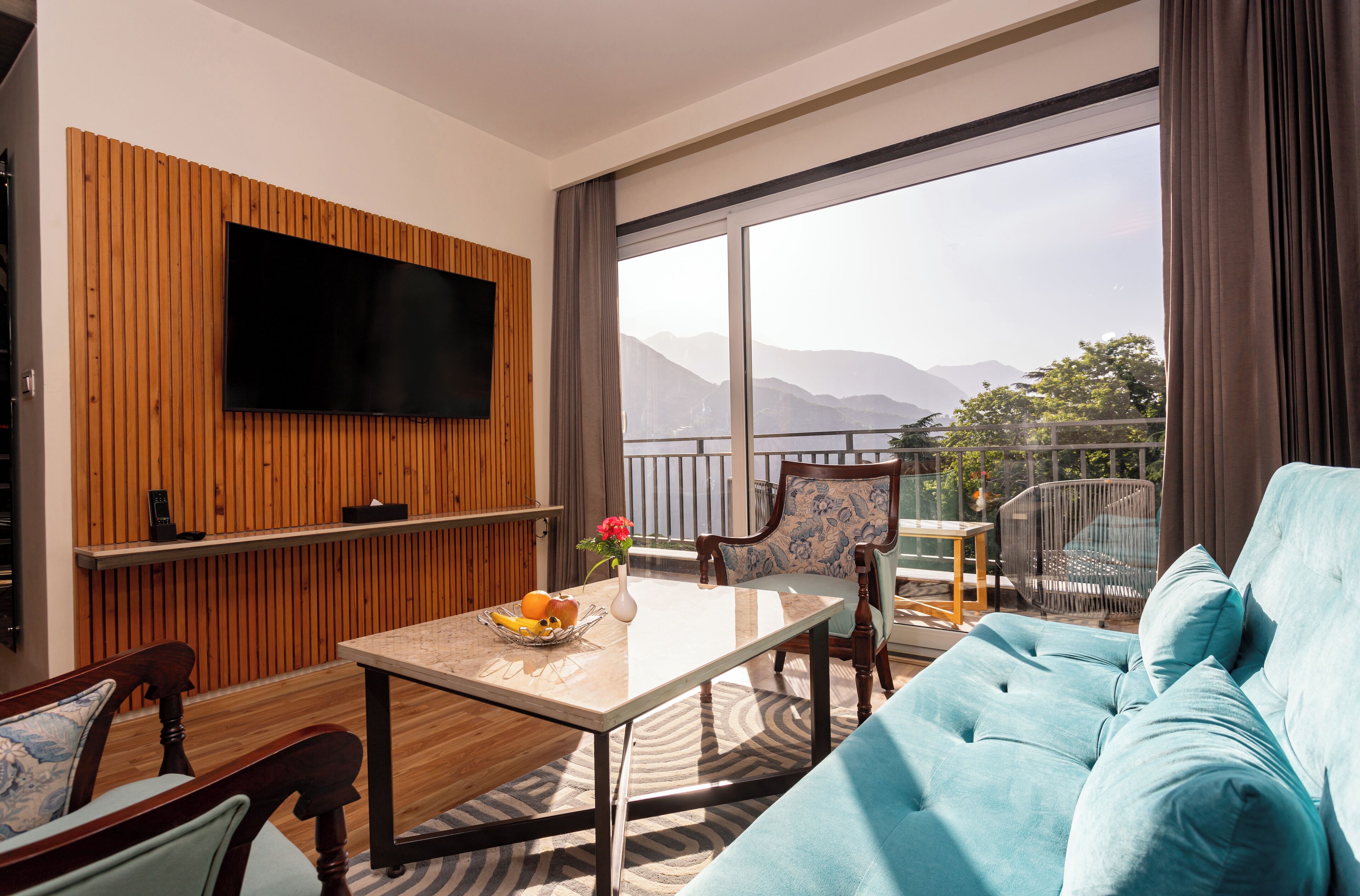 A modern living room with a flat-screen TV, wooden panel wall, a sofa, a dining table with fruit, and a balcony with a view of mountains.