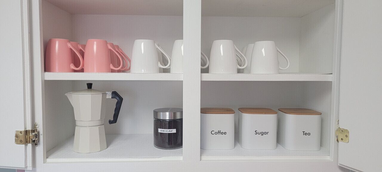 Un armario de cocina con tazas rosas y blancas, una cafetera y recipientes etiquetados para café, azúcar y té.