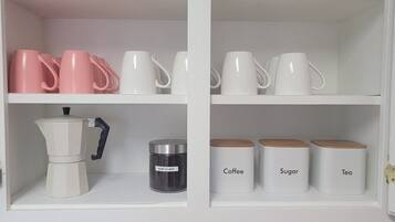 Un armario de cocina con tazas rosas y blancas, una cafetera y recipientes etiquetados para café, azúcar y té.