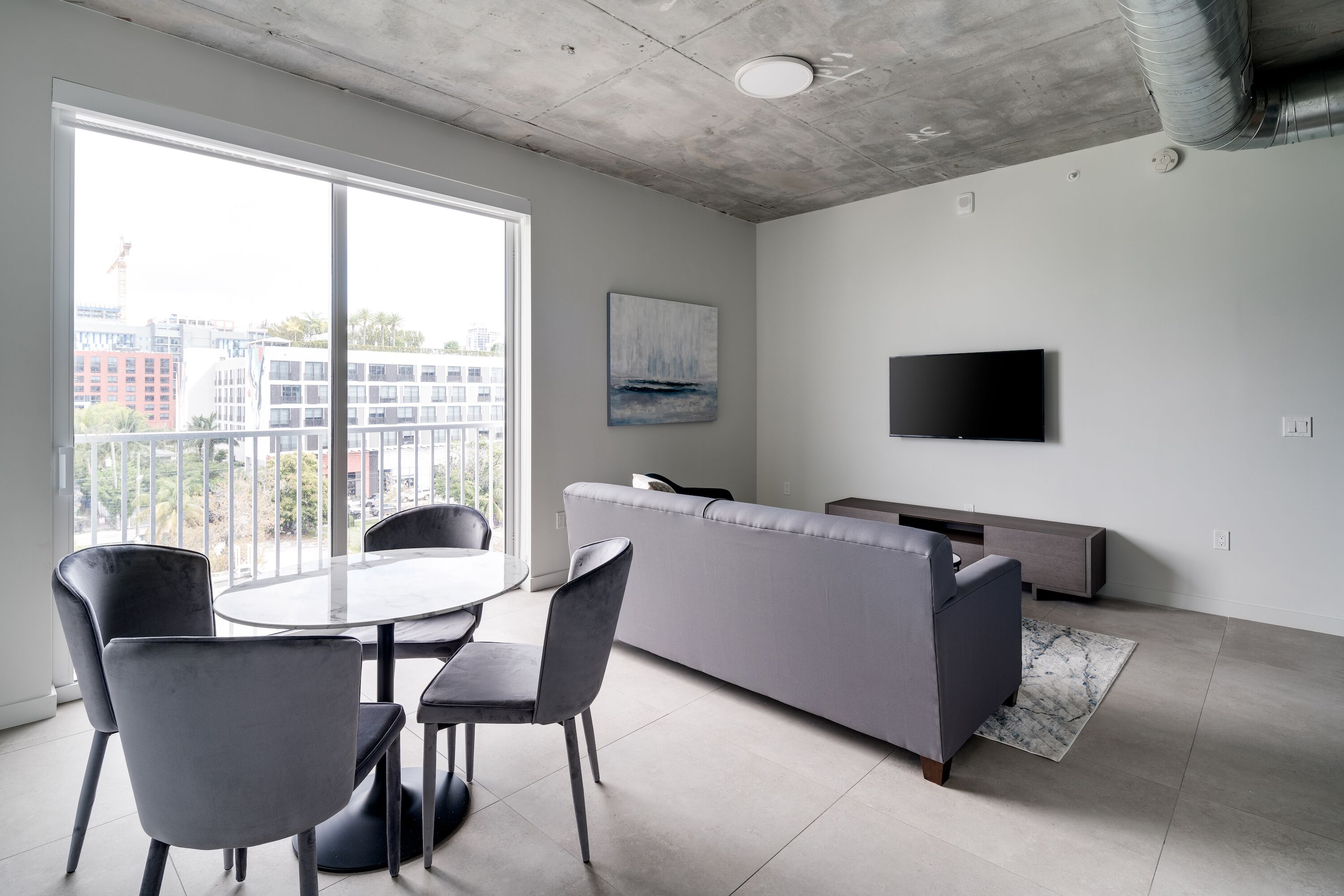 Een moderne woonkamer met een grijze bank, een ronde eettafel met stoelen, een flatscreen-tv aan de muur en grote ramen met uitzicht op gebouwen.