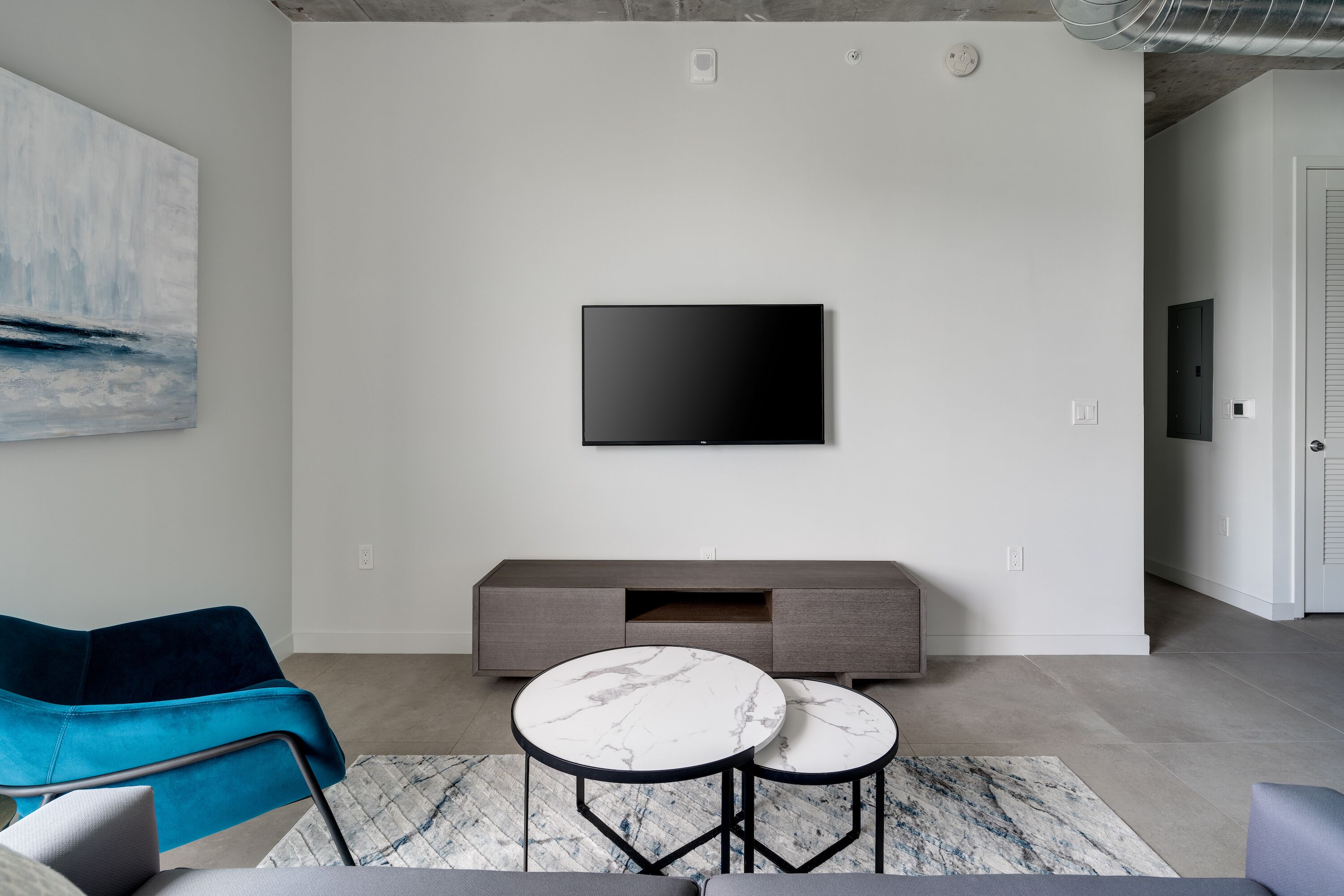 A modern living room with a flat-screen TV mounted on the wall, a grey sofa, and two round tables with marble tops.