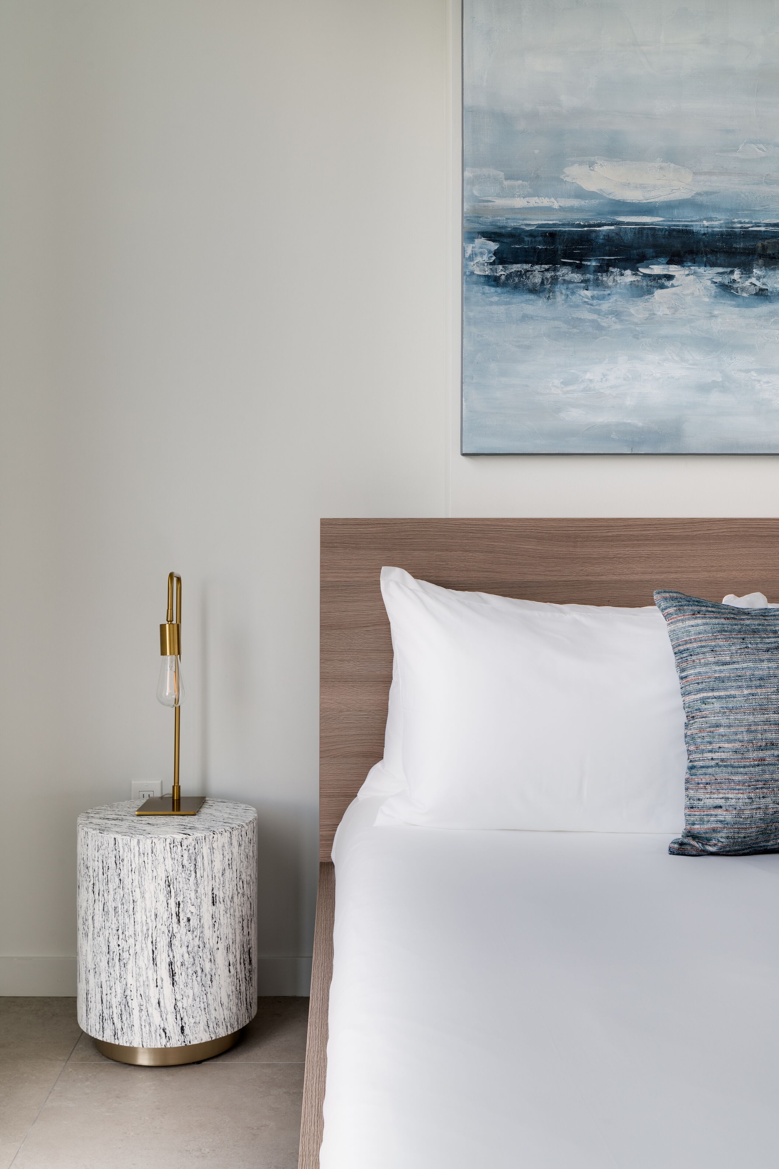 A modern hotel room with a wooden headboard, a white bed with a blue and white patterned pillow, and a marble side table with a gold lamp.