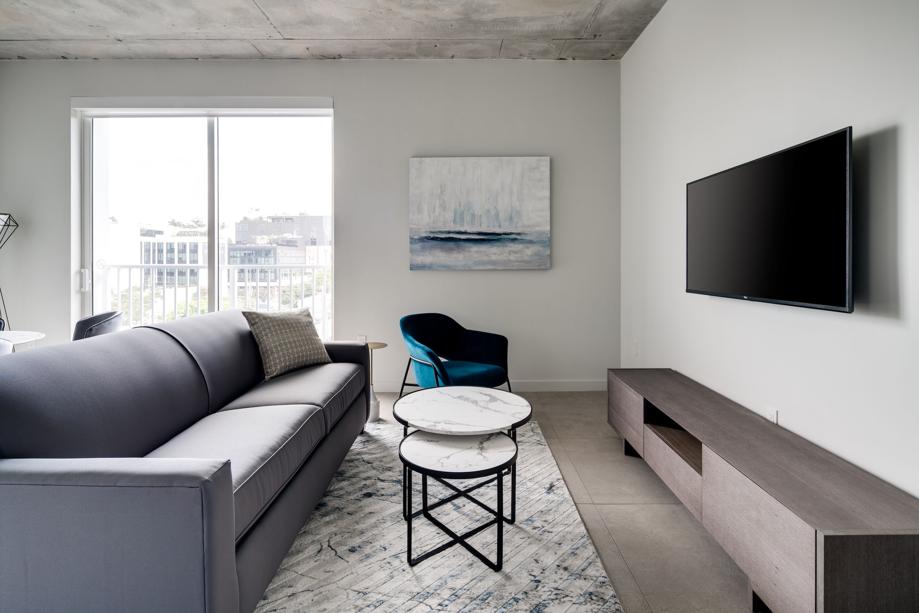 Una sala moderna con un sofá gris, un sillón azul, una mesa de centro de mármol y un televisor de pantalla plana montado en la pared.