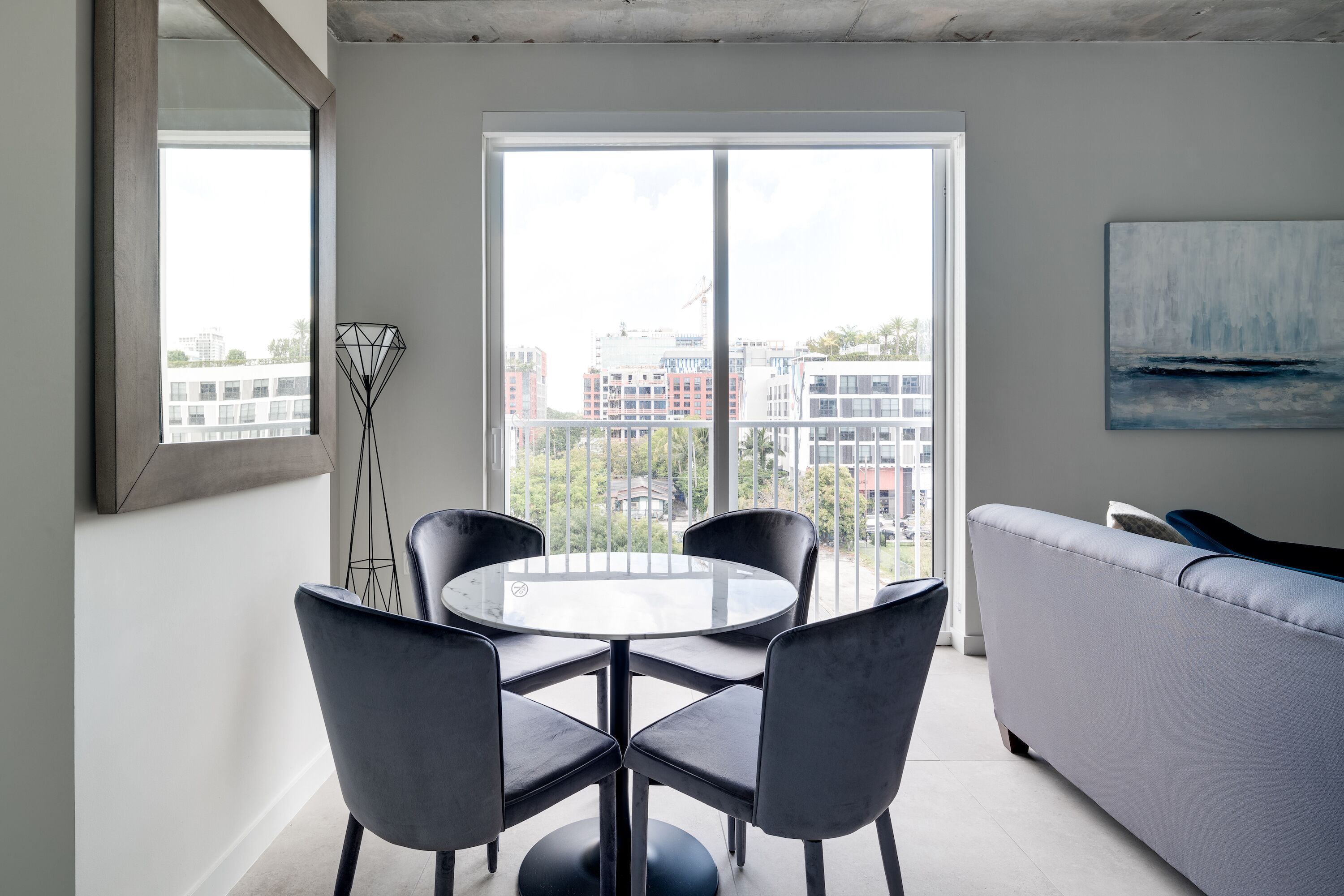 A modern living room with a round glass table surrounded by four chairs, a grey sofa, and a large window offering a city view.
