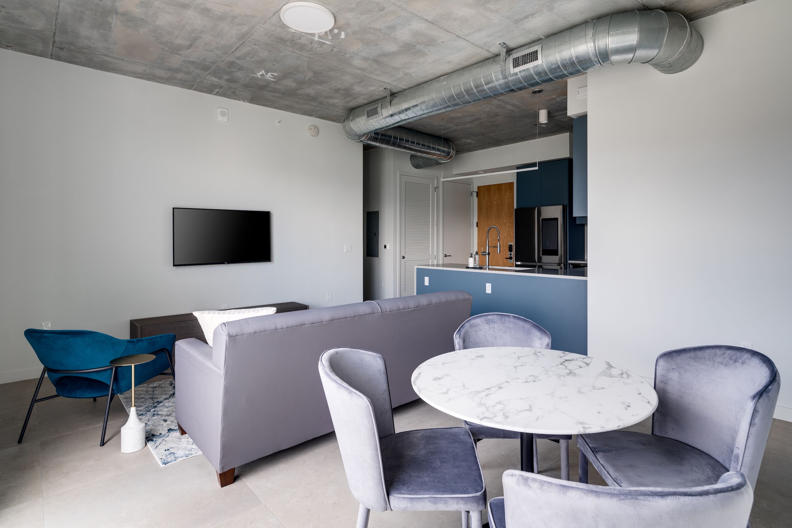 A modern living room with a grey sofa, a round marble table, and matching chairs. A flat-screen TV is mounted on the wall, and a kitchen area is visible in the background.