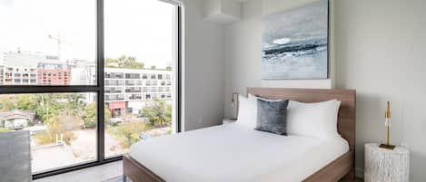 Quarto moderno com uma grande janela, cama com roupa de cama branca, cabeceira de madeira, mesa de cabeceira redonda e vista para a cidade.