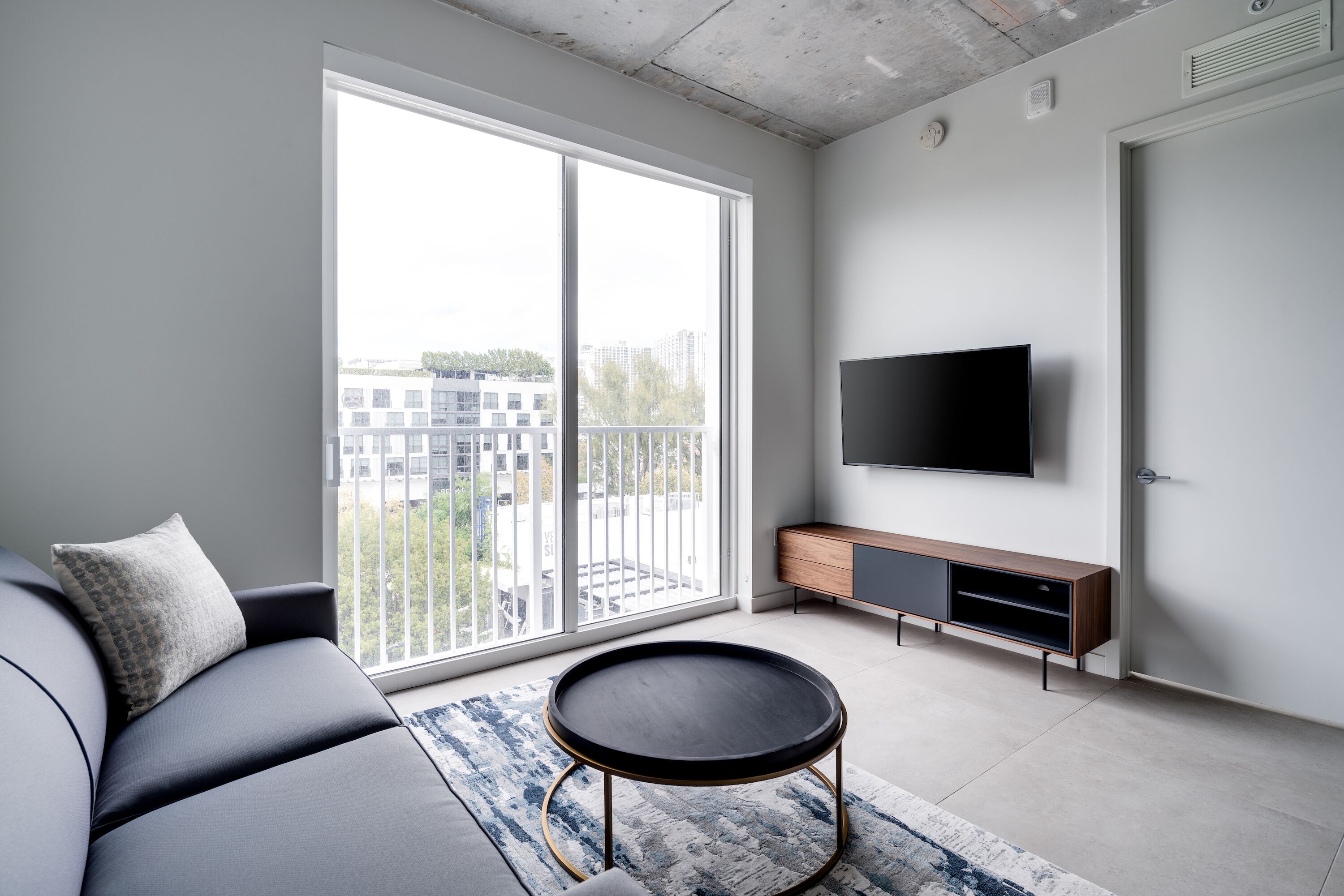 Un salón moderno con un sofá, una mesa redonda de centro, un televisor y una puerta corrediza de vidrio a un balcón.