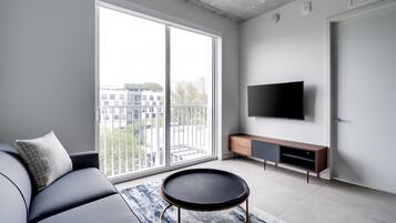 Un salon moderne avec un canapé, une table basse ronde, un téléviseur et une porte-fenêtre coulissante donnant sur un balcon.