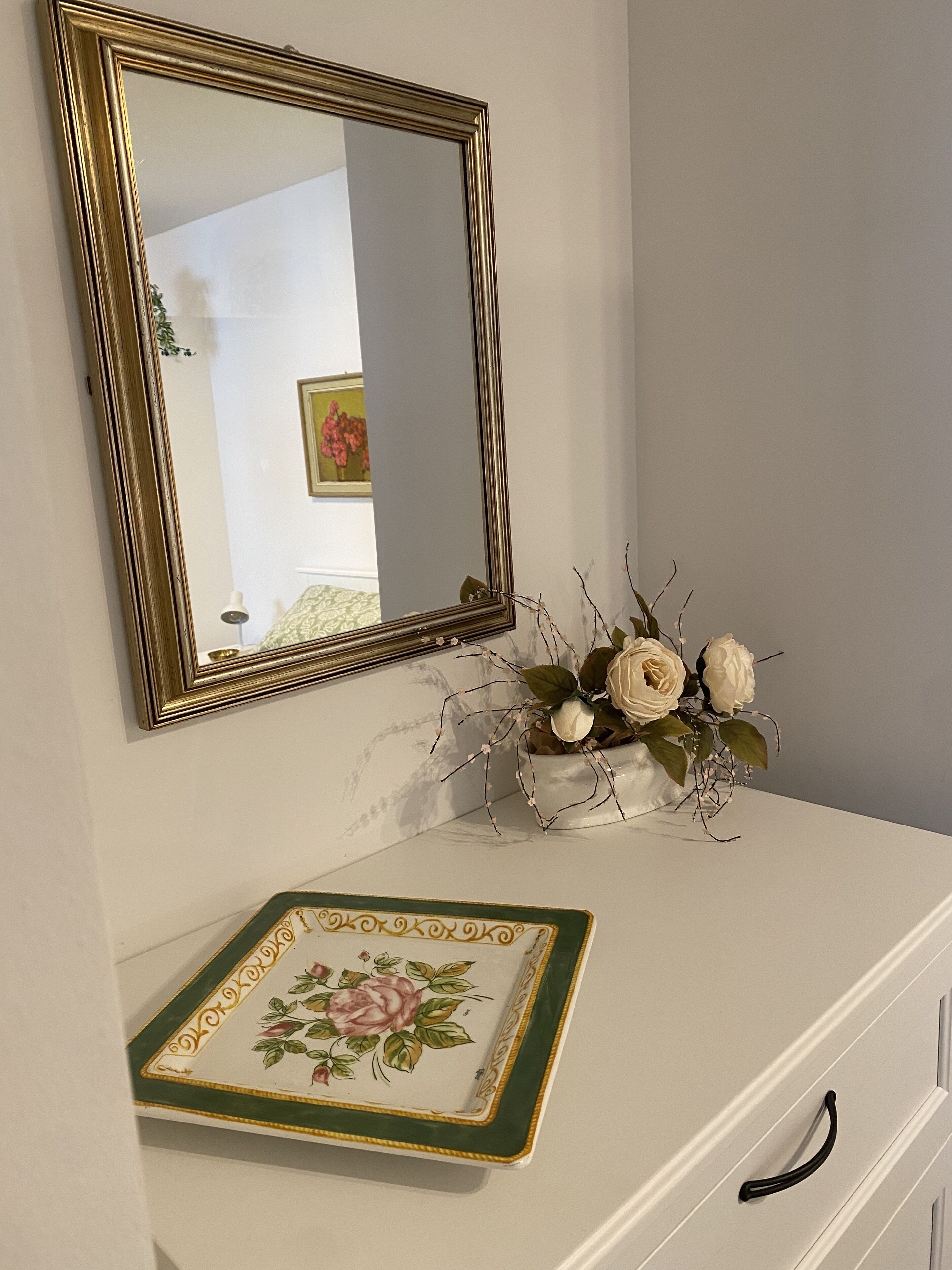 A white dresser with a gold-framed mirror above it, a decorative plate with floral designs, and a vase with white flowers.