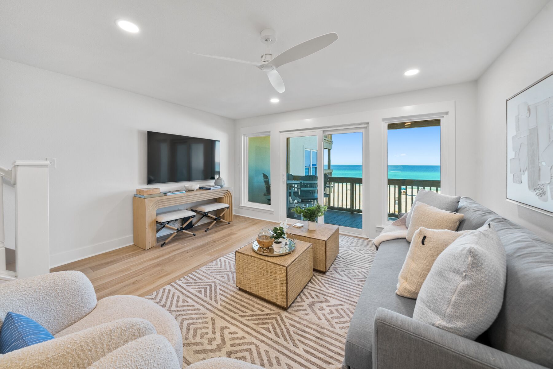 A modern living room with a flat-screen TV, a wooden desk, a sofa, and a balcony with a view of the beach.