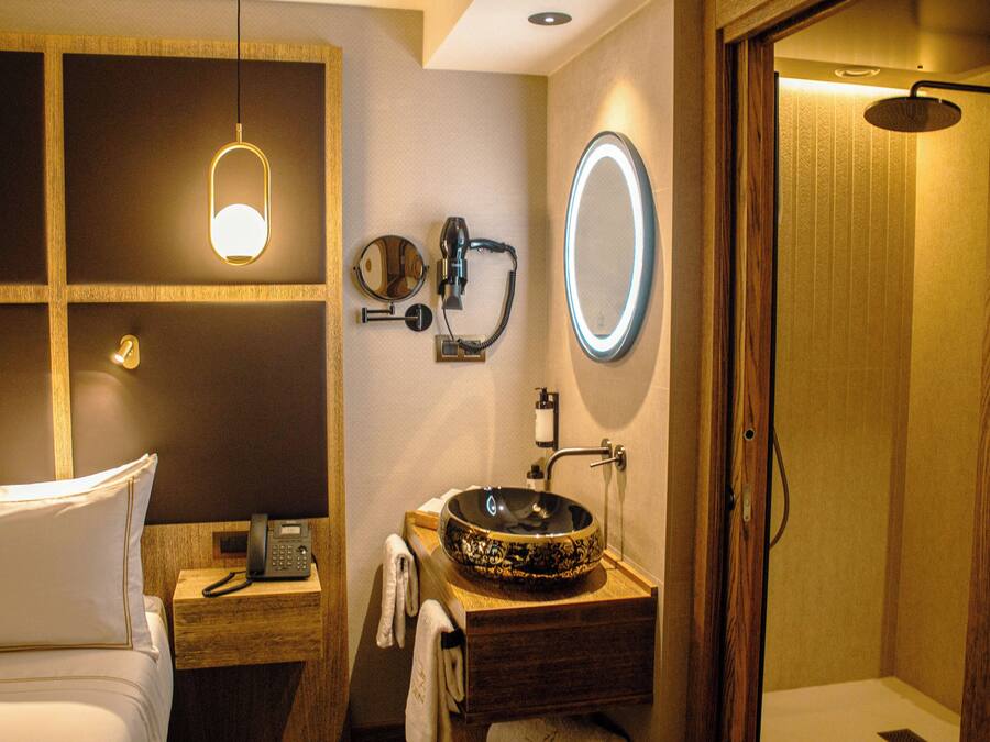 A hotel bathroom with a wooden vanity, a round mirror, a built-in sink, a handheld showerhead, and a bedside telephone.