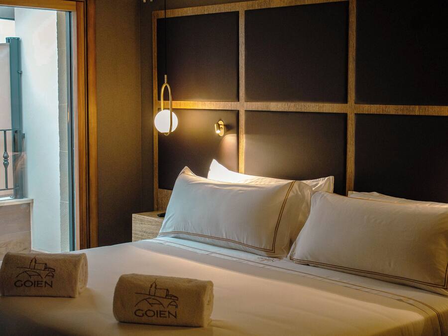 Habitación de hotel con una cama grande, dos almohadas, una mesita de noche, una lámpara y dos toallas con el nombre del hotel.